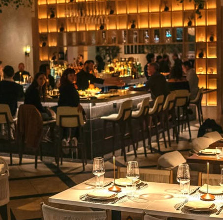 Cozy upscale city restaurant and cocktail bar with warm backlit shelves, patrons seated at a curved marble bar, and candlelit tables set for dinner in the foreground
