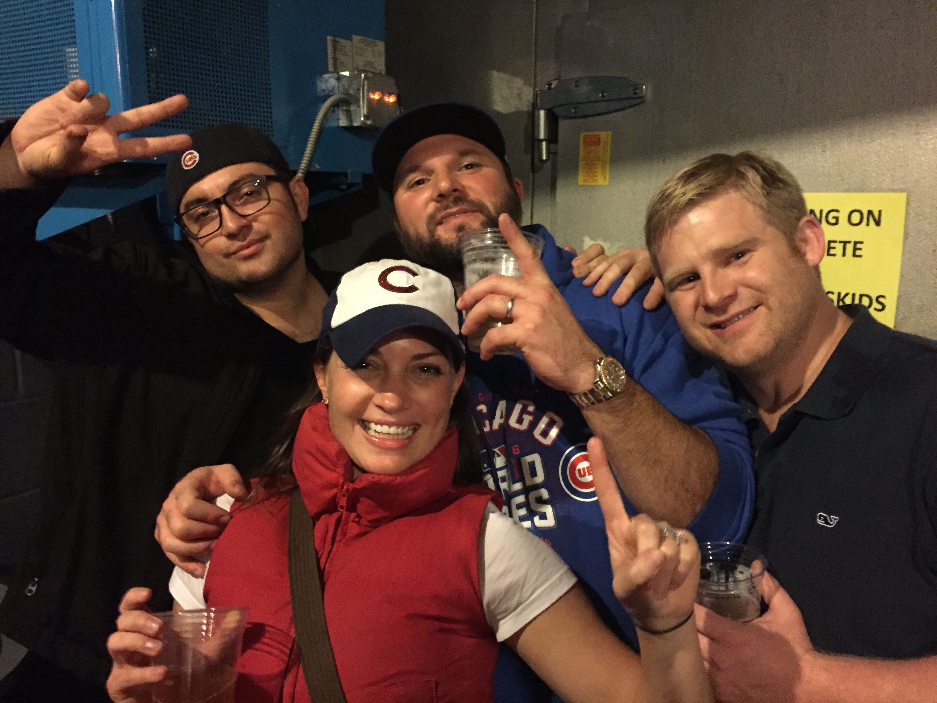Four smiling Chicago Cubs fans in caps and a team hoodie, holding plastic beers and striking playful poses in a stadium concourse.
