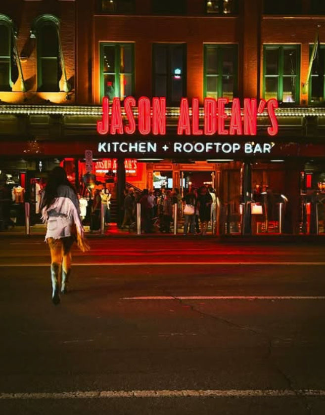 Vibrant neon-lit downtown rooftop bar and kitchen entrance at night, crowds inside and a lone pedestrian crossing the street.