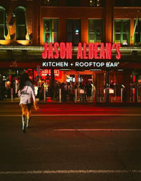 Vibrant neon-lit downtown rooftop bar and kitchen entrance at night, crowds inside and a lone pedestrian crossing the street.