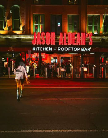 Vibrant neon-lit downtown rooftop bar and kitchen entrance at night, crowds inside and a lone pedestrian crossing the street.