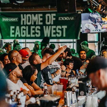 Lively sports bar crowd packed at the bar, fans in green jerseys cheering under a large fan banner while a football game plays on TV