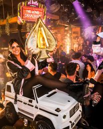 Smiling woman in a satin robe sits on a white toy SUV in a packed nightclub, holding a gold spade-shaped champagne display amid confetti, sparklers, neon lights and a lively crowd — energetic VIP bottle-service party scene.
