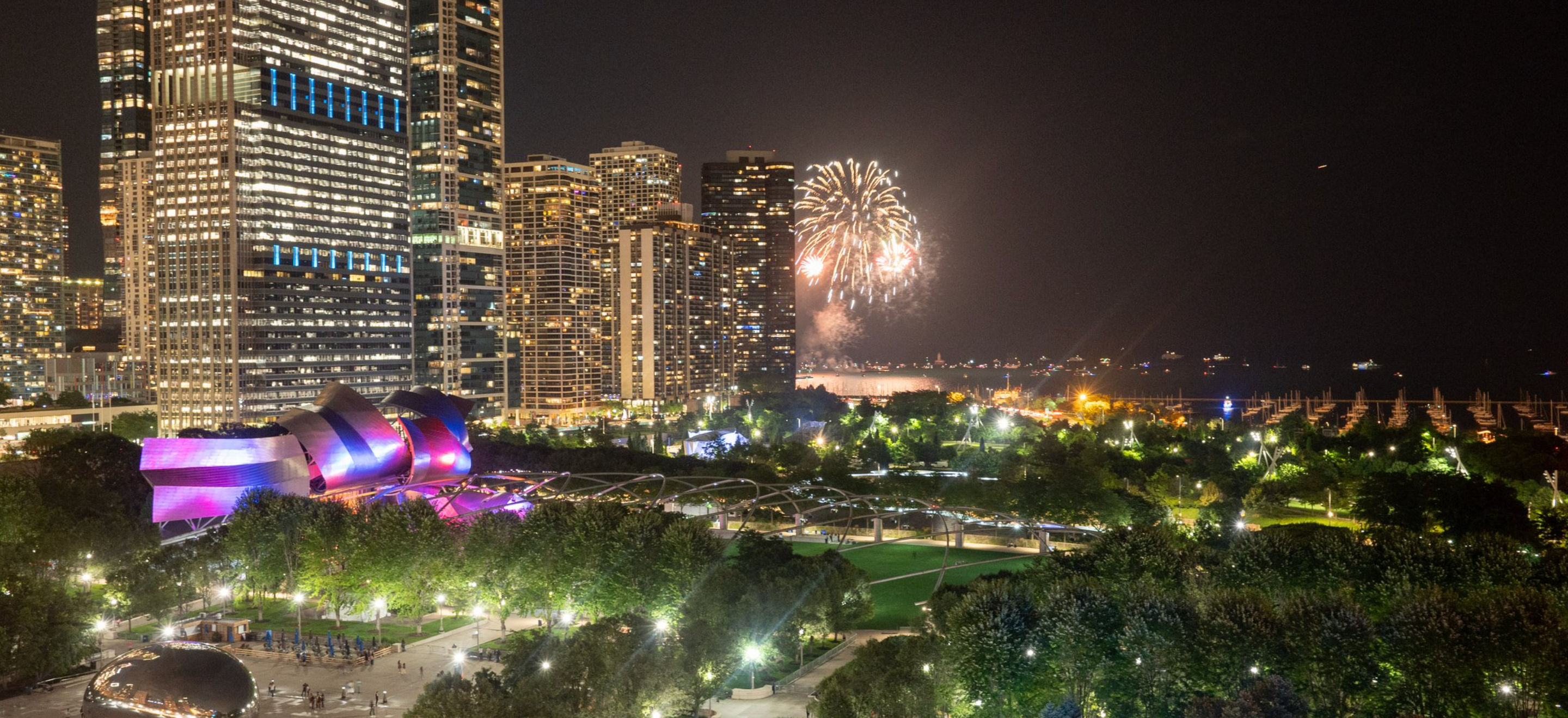 Chicago skyline at night over a lakefront park — illuminated modern bandshell and reflective public sculpture near lit walking paths and trees, with fireworks bursting over the water