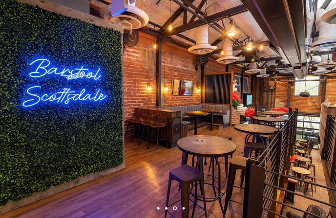 Industrial‑chic Scottsdale bar lounge with a neon cursive sign on a greenery wall, exposed brick, wooden floors, high-top tables and cozy booths under warm pendant lighting