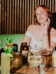 Woman in an off-shoulder top smiling at a cocktail bar, surrounded by mixed drinks, a beer bottle and mint-garnished cocktails on a wooden table with corrugated wood backdrop.