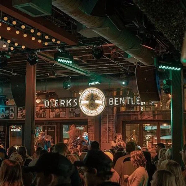Packed indoor country-music venue with a live band on stage beneath a glowing circular logo and marquee, warm string lights, exposed ceiling ducts, and an energetic nightlife crowd.
