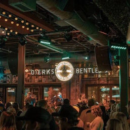 Packed indoor country-music venue with a live band on stage beneath a glowing circular logo and marquee, warm string lights, exposed ceiling ducts, and an energetic nightlife crowd.