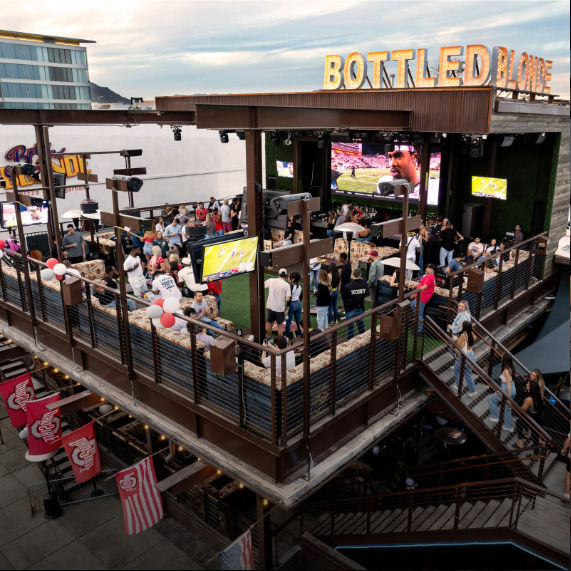 Packed downtown rooftop bar patio with multi-level wooden deck, crowds watching a football game on large outdoor screens, string lights and festive balloons, urban nightlife vibe.