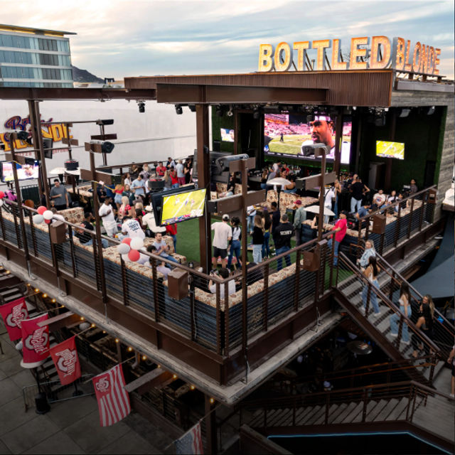 Packed downtown rooftop bar patio with multi-level wooden deck, crowds watching a football game on large outdoor screens, string lights and festive balloons, urban nightlife vibe.