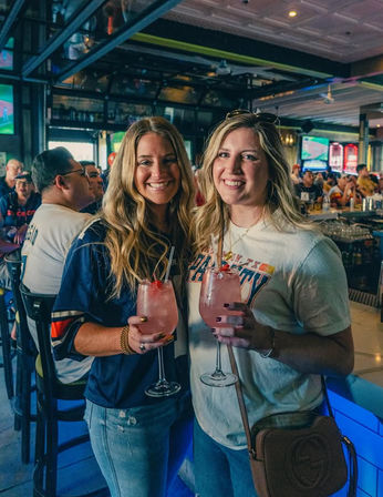 Two smiling friends holding pink cocktails in stemmed glasses at a lively neon-lit sports bar, TVs and a crowded bar counter visible in the background.