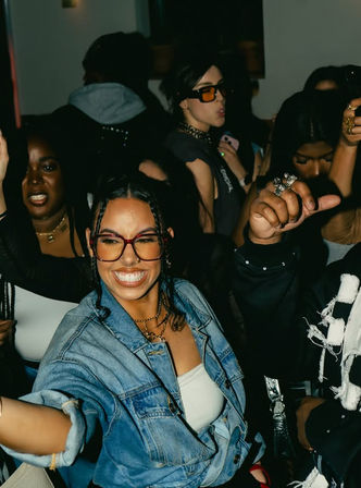 Smiling woman in denim jacket and glasses dancing in a packed indoor nightlife party, hands raised and friends around her.