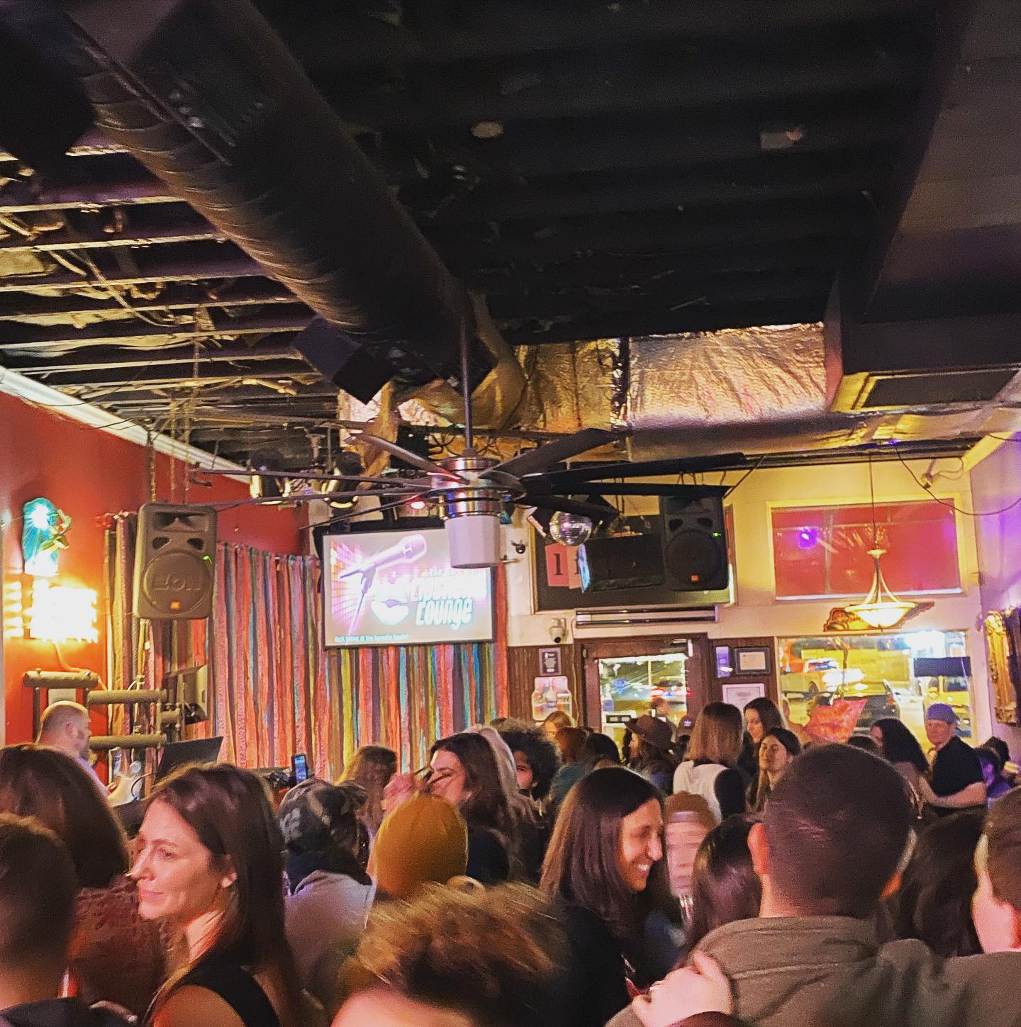 Bustling bar lounge interior at night with a crowd socializing under exposed ceiling ducts and a large fan, colorful stage curtains, neon lights and speakers.
