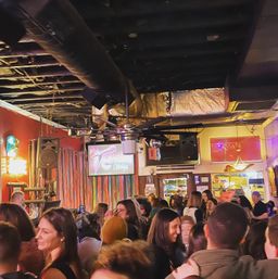 Bustling bar lounge interior at night with a crowd socializing under exposed ceiling ducts and a large fan, colorful stage curtains, neon lights and speakers.