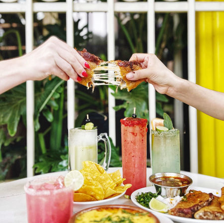 Hands pulling apart a gooey cheese quesadilla above a table with tortilla chips, salsa, grilled meat and three colorful fruit cocktails on a sunny outdoor restaurant patio.