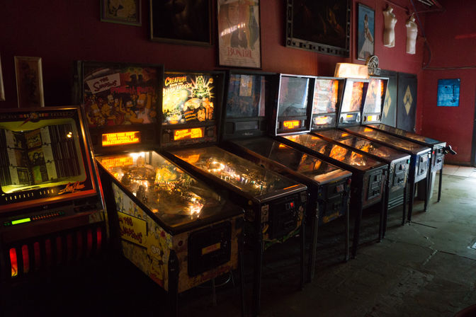 Dimly lit retro arcade corner with a row of vintage illuminated pinball machines against a red wall, nostalgic gaming bar vibe