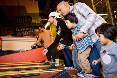 Family playing skee-ball at a bustling indoor arcade: a father guiding a young child as others roll balls down wooden lanes under warm lights.