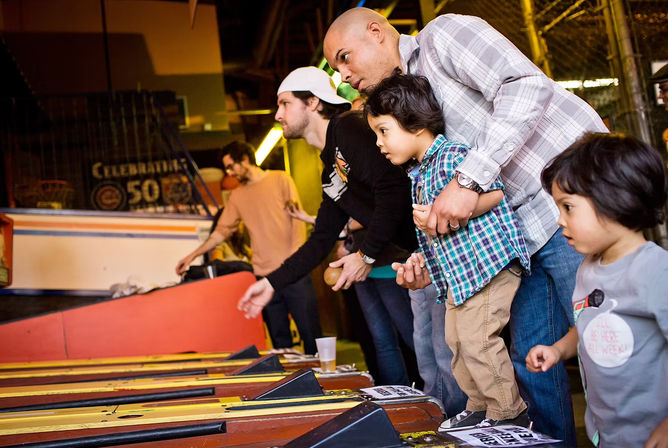 Family playing skee-ball at a bustling indoor arcade: a father guiding a young child as others roll balls down wooden lanes under warm lights.
