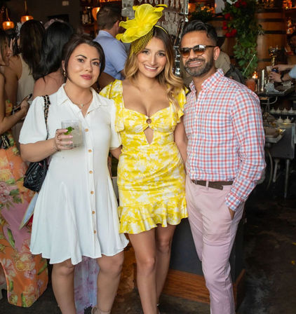 Three people smiling inside a busy bar: central woman in a bright yellow floral mini dress with matching fascinator, flanked by a woman in a white button-front dress holding a cocktail and a man in sunglasses wearing a pink plaid shirt and light trousers.
