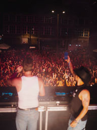 Two DJs on stage hyping a packed nighttime downtown crowd at an outdoor music festival, sea of raised hands and glowing phones under warm stage lights