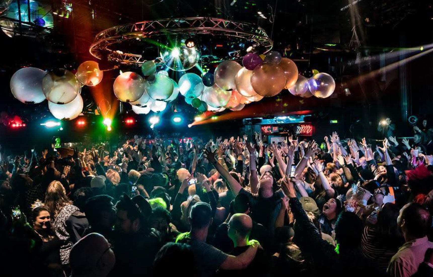 Packed nightclub dance floor with cheering crowd under disco balls and colorful stage lights