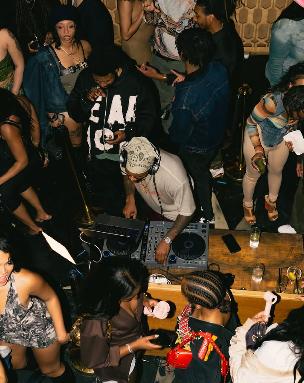 Overhead view of a packed nightclub DJ booth — DJ wearing headphones mixing on turntables as partygoers dance, mingle and sip drinks at the bar.