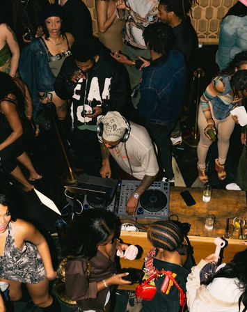 Overhead view of a packed nightclub DJ booth — DJ wearing headphones mixing on turntables as partygoers dance, mingle and sip drinks at the bar.