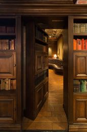 Wood-paneled library with a secret bookcase door opening to a cozy leather chesterfield reading nook lit by a warm lamp, antique books and hardwood floors.