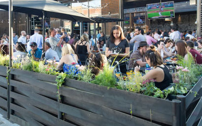 Busy urban outdoor patio beer garden with wooden planters and flowering greenery, diners and friends socializing under umbrellas while a server takes orders and TVs show a baseball game.