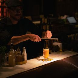 Bartender grating citrus zest over a steaming orange-hued cocktail in a footed glass on a marble bar, dimly lit speakeasy-style cocktail bar scene