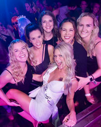Smiling group of women at a neon-lit nightclub celebrating a bachelorette party, bride-to-be in a white dress and sash surrounded by friends in black outfits.