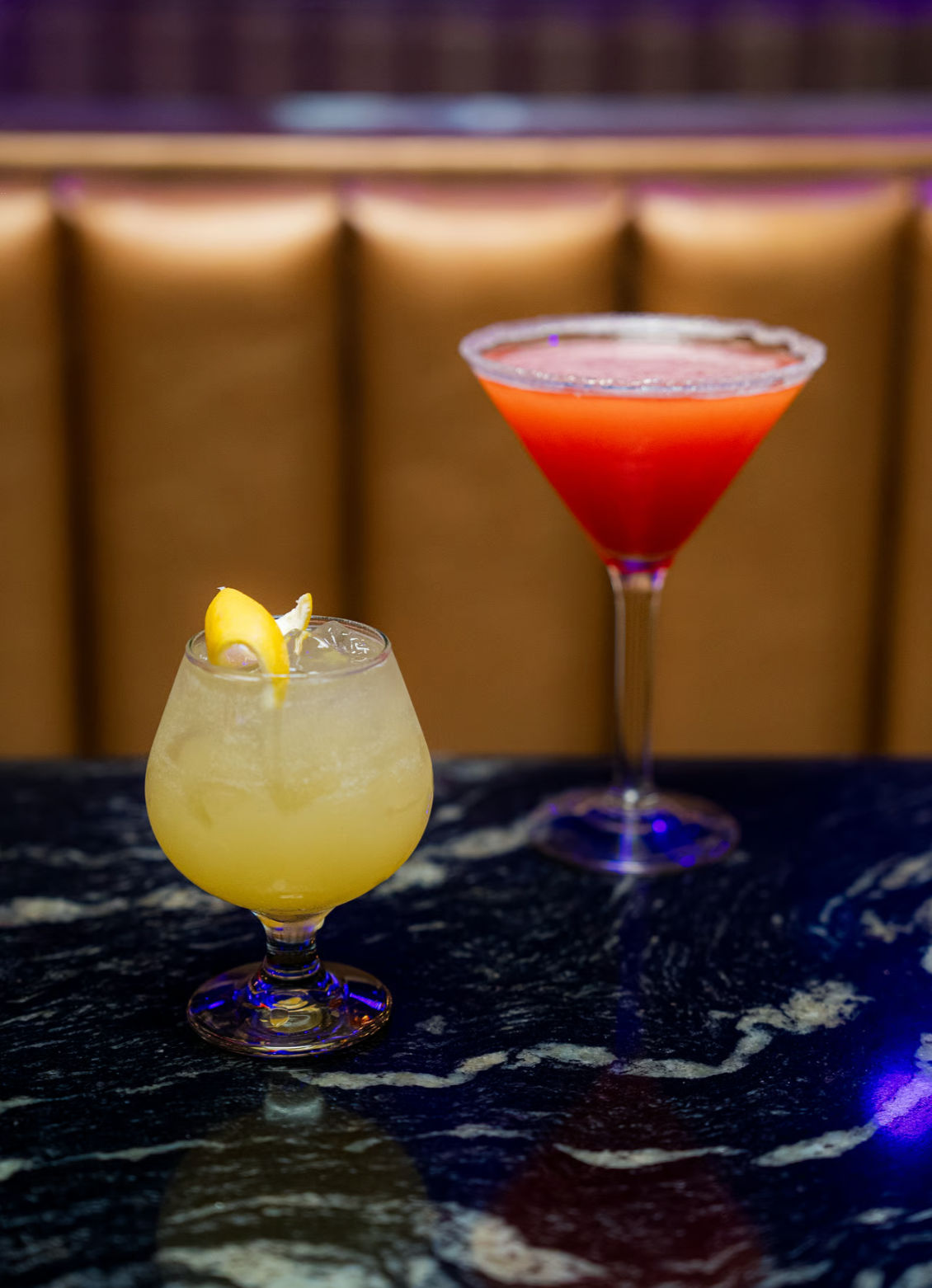 Two colorful cocktails on a dark marble bar countertop: a frosty yellow citrus drink with a lemon twist in a stemmed snifter and a bright red martini with a sugared rim in a martini glass.
