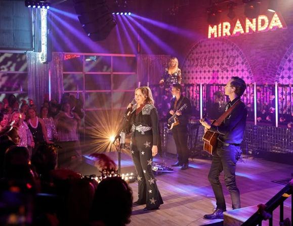 Female lead singer in a star-covered black jumpsuit sings into a microphone with a guitarist and band on a colorful, marquee-lit stage while an enthusiastic crowd watches.