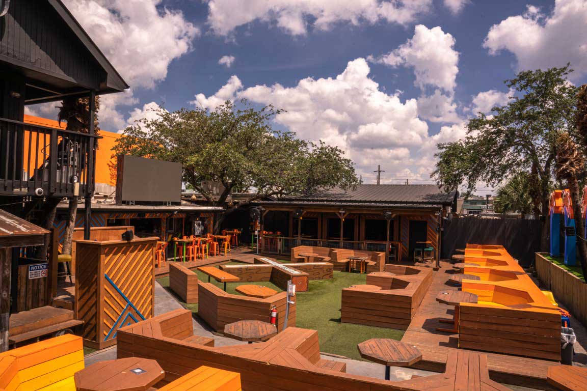 Sunlit urban courtyard patio with geometric wooden seating, bright orange benches and high bar tables beneath leafy trees and puffy clouds.