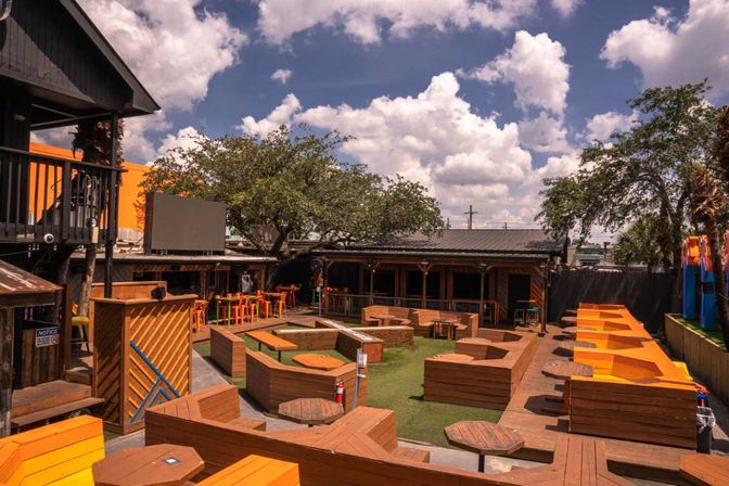 Sunlit urban courtyard patio with geometric wooden seating, bright orange benches and high bar tables beneath leafy trees and puffy clouds.