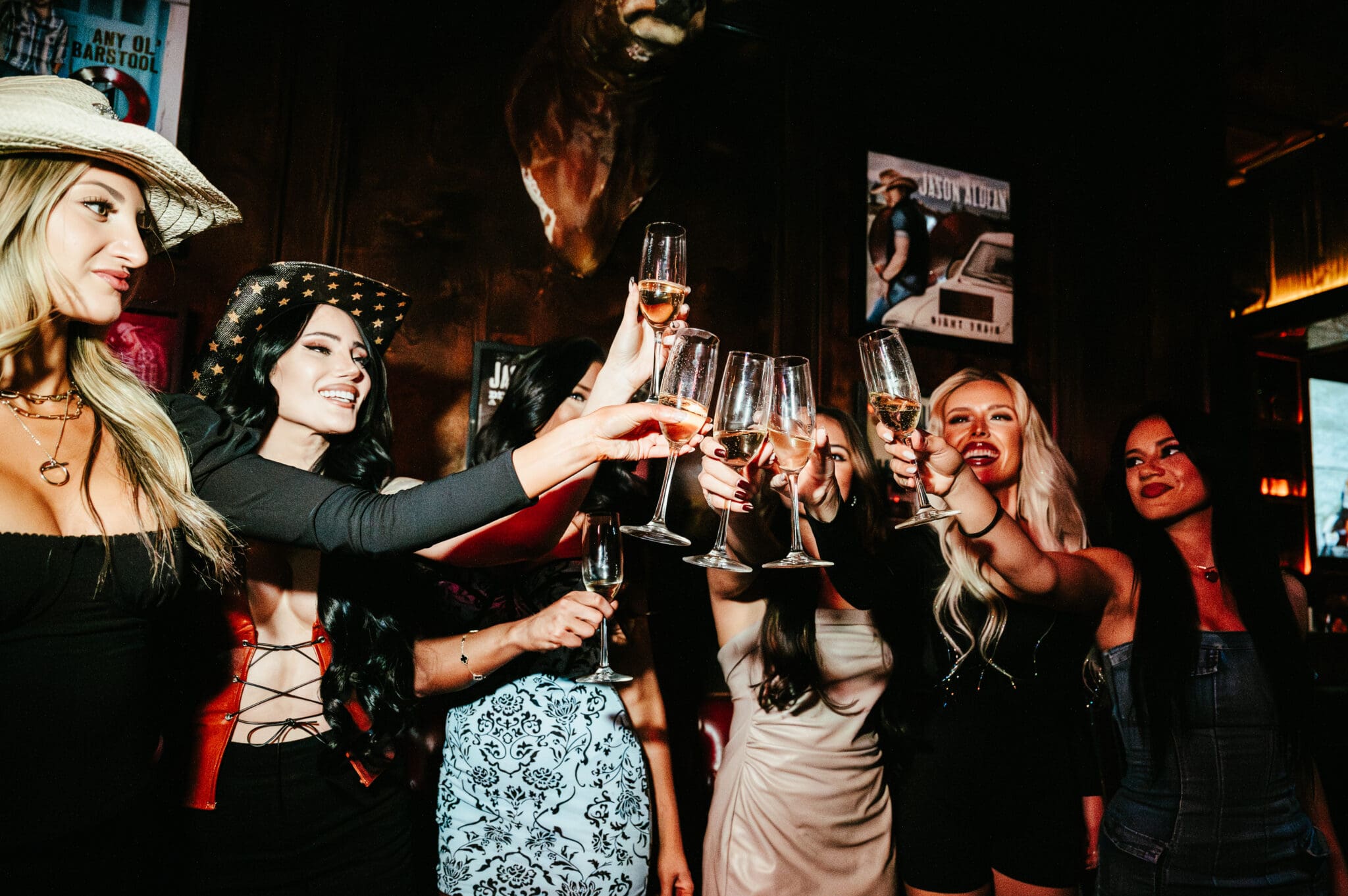 Group of women toasting with champagne flutes in a dim, festive bar interior — stylish outfits and cowboy hats creating a lively girls' night out vibe.