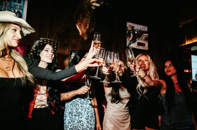 Group of women toasting with champagne flutes in a dim, festive bar interior — stylish outfits and cowboy hats creating a lively girls' night out vibe.