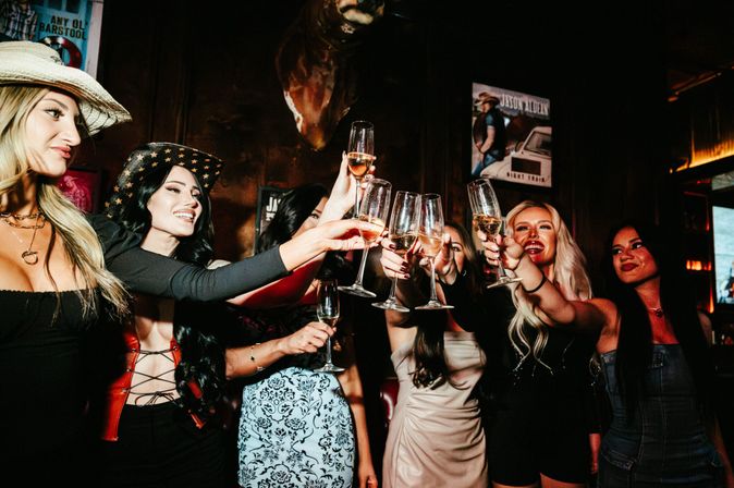 Group of women toasting with champagne flutes in a dim, festive bar interior — stylish outfits and cowboy hats creating a lively girls' night out vibe.