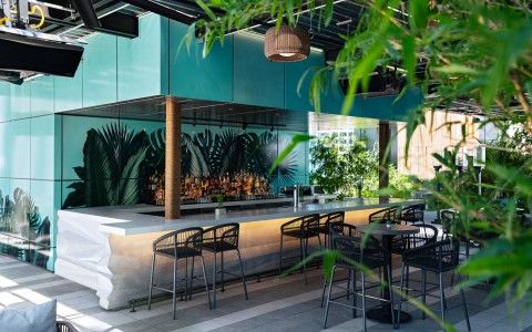 Modern rooftop bar patio with a long backlit white counter, black rattan barstools, hanging wicker lights and lush tropical plants in a bright urban outdoor lounge.