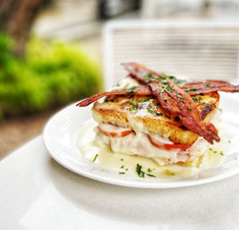 Croque-monsieur style brunch sandwich with toasted bread, melted cheese and béchamel, tomato and ham layers, topped with crispy bacon and parsley on a white plate outdoors