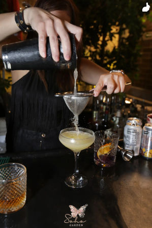 Bartender pouring a shaken citrus cocktail through a fine mesh strainer into a coupe glass on a bustling urban bar counter, with mixing glass, garnish cup and cans nearby.
