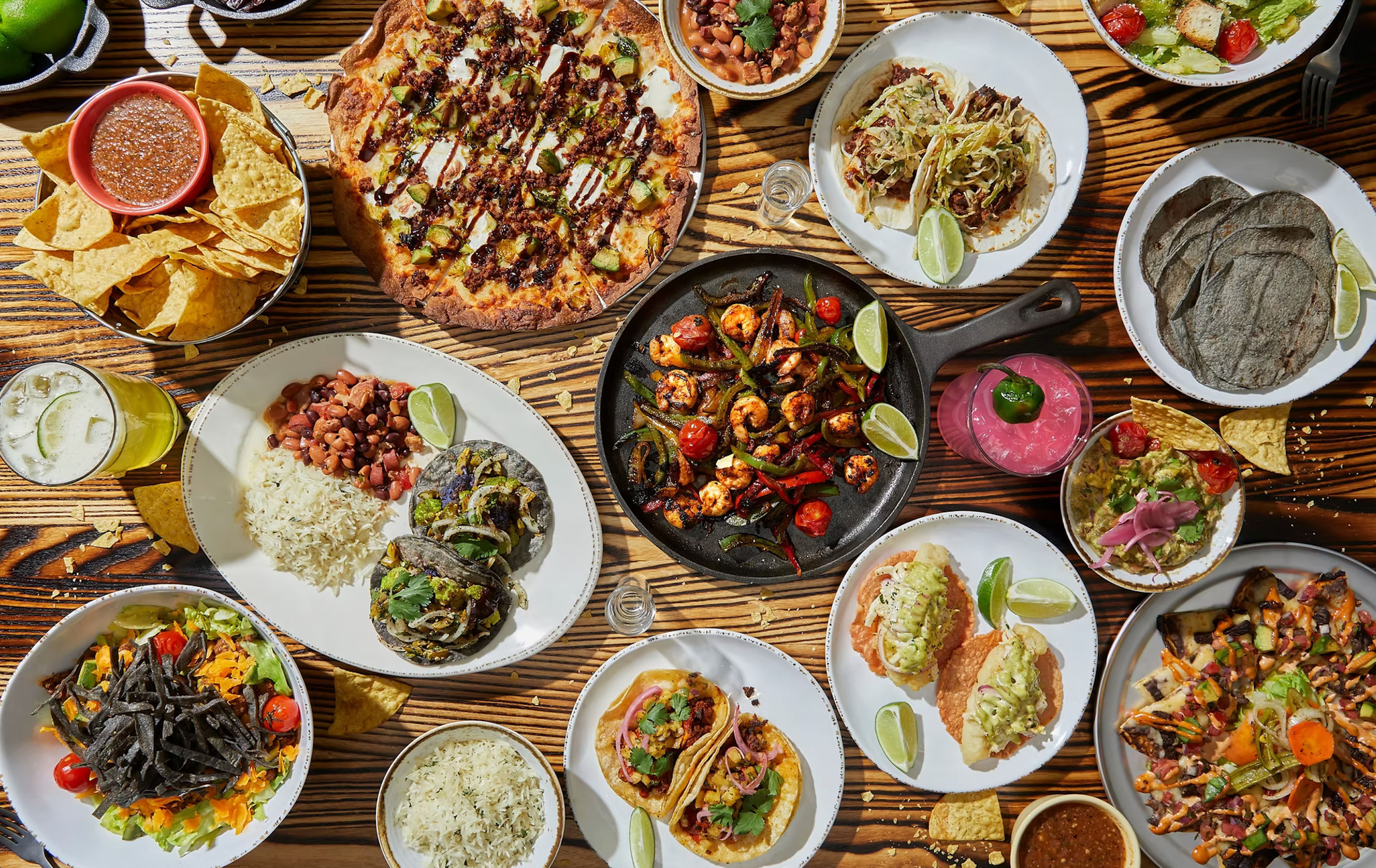 Overhead shot of a vibrant Mexican-style spread on a wooden table, featuring tacos, grilled shrimp fajitas in a cast-iron skillet, chips and salsa, guacamole, rice and beans, blue corn tortillas and colorful cocktails.