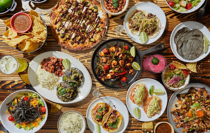 Overhead shot of a vibrant Mexican-style spread on a wooden table, featuring tacos, grilled shrimp fajitas in a cast-iron skillet, chips and salsa, guacamole, rice and beans, blue corn tortillas and colorful cocktails.
