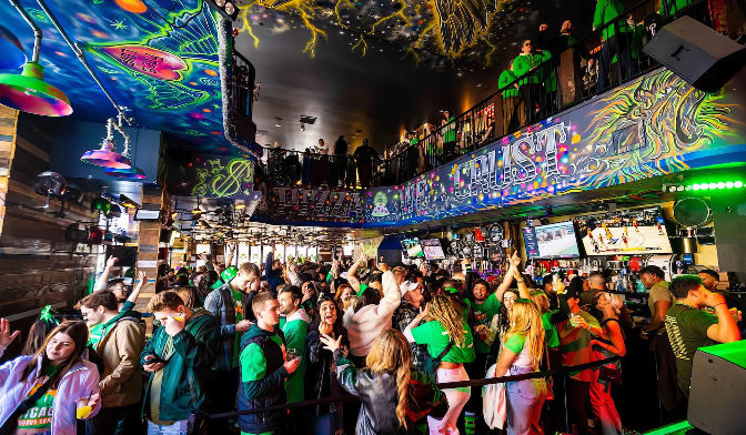 Vibrant neon-lit bar interior with balcony and psychedelic ceiling art; crowded patrons in green celebrating and watching sports on TVs.