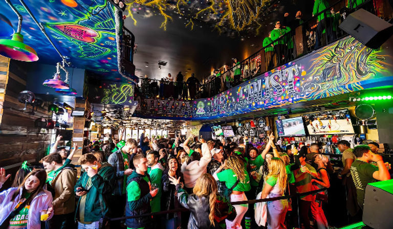 Vibrant neon-lit bar interior with balcony and psychedelic ceiling art; crowded patrons in green celebrating and watching sports on TVs.