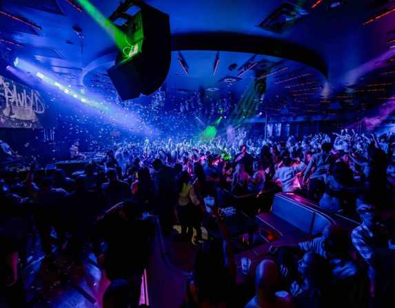 Packed downtown nightclub dance floor lit in blue and purple, DJ booth in the background, confetti and green laser beams over a cheering crowd