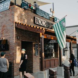 Brick sports bar exterior with wooden awning and rooftop patio, green-and-white fan flags and banners, people on the sidewalk on a sunny afternoon.