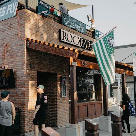Brick sports bar exterior with wooden awning and rooftop patio, green-and-white fan flags and banners, people on the sidewalk on a sunny afternoon.