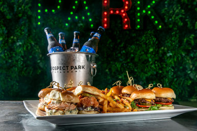 Assorted mini burgers and crispy seasoned fries on a rectangular platter with an ice bucket of bottled beer, set against a leafy wall lit with green and red lights — casual bar food scene.