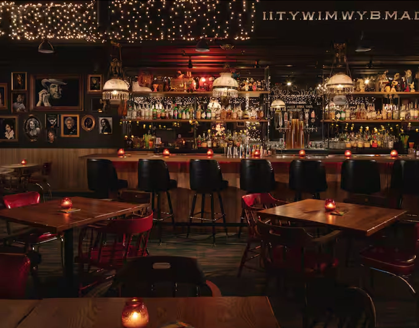 Cozy dimly lit cocktail bar interior with wooden tables and red votive candles, black barstools at a long wood bar stocked with bottles, vintage pendant lamps, string fairy lights overhead and framed portraits on the wall — warm, intimate urban vibe.